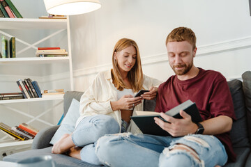 Image of ginger happy couple using cellphone and reading book