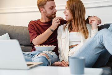 Image of joyful couple eating popcorn while watching movie on laptop