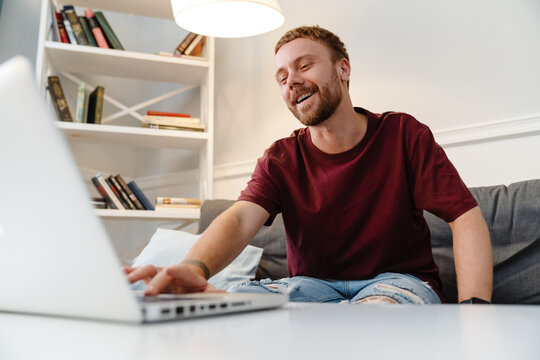 Image of ginger guy laughing and using laptop while sitting on sofa