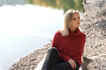 beautiful young blonde woman sitting by the lake in a red sweater. autumn concept