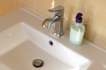 View of a white bathroom sink with a contemporary mixer tap, an unlabeled bottle of liquid soap. Concept of modern interior, water saving, everyday hygiene. Side view, close-up. Horizontal format