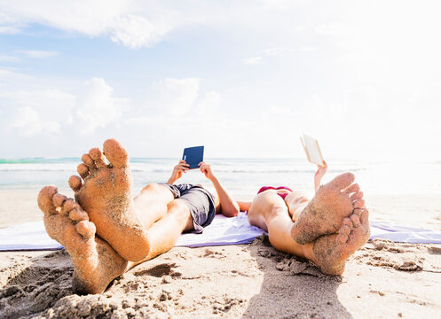 Close-up of sand-covered feet of young couple lying on blanket on beach reading