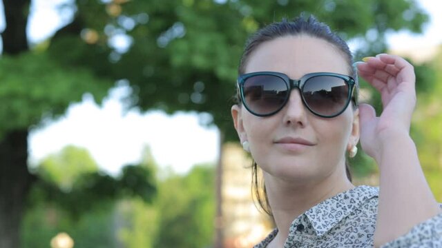 Portrait of a young woman with dark hair in a blouse, blue eyes and sunglasses on the background of a Park. She turns her head,takes off her glasses, and looks into the distance, smiling and dreaming.