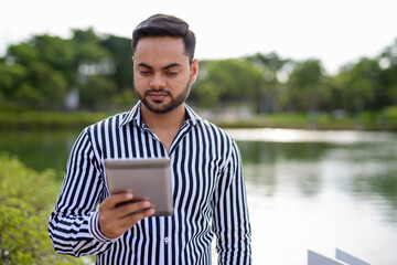 Fototapeta premium Young bearded Indian businessman relaxing at the park in the cit