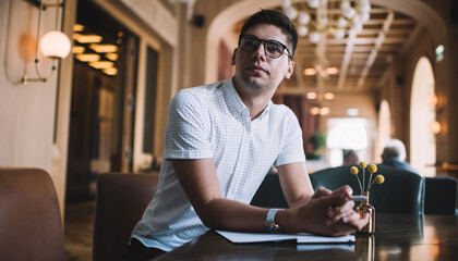 Serious young man with pen and notebook sitting in cafe with hands clasped