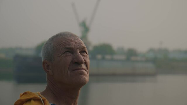 Close-up: portrait of a thoughtful adult man in a yellow raincoat, outdoors by the river, looking up into the distance. Gray-haired old man with wrinkles in nature in the setting sun. Slow motion. 4K