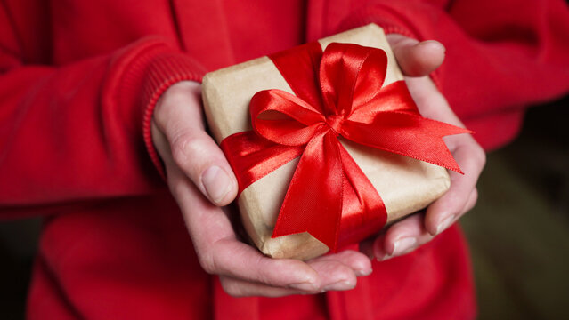 Young woman in red swether holding a gift box wrapped in brown paper and tied with a red ribbon with a bow for a space event. Christmas, New Year, Birthday, Valentine's Day.Close-up