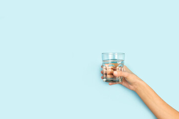 Mano de mujer sosteniendo un vaso con agua sobre un fondo celeste liso y aislado. Vista de frente. Copy space