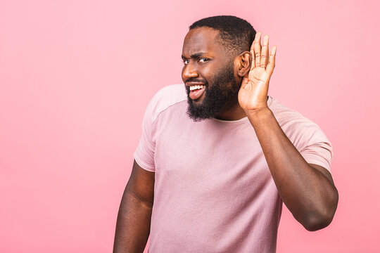 African American Man With Beard Holding Hand Near Ear Trying To Listen To Interesting News Expressing Communication Concept And Gossip Isolated Over Pink Background.