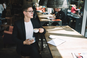 Thoughtful young man using smartphone and drinking coffee in cafeteria