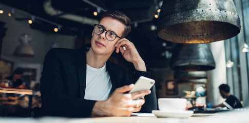 Pensive male cafe worker with smartphone and cup of coffee