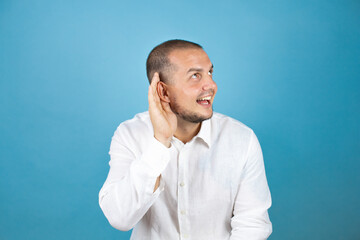 Russian business man wearing white shirt standing over blue background smiling with hand over ear listening an hearing a rumor or gossip. deafness