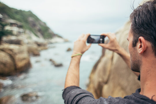 Young man taking photos with his phone with rocky coastline and water in background