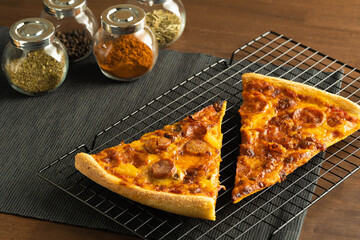 Two pizza with pepperoni topping on a cooling rack place on black cloth placemat and a group of herbs and spices in jars on a conners