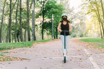 Young beautiful woman wearing protective mask against coronavirus riding an electric scooter in a city park- concept of sustainable personal mobility