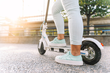 close up of young woman legs riding an electric scooter on the asphalt in a city at sunset with blurred background - concept of sustainable personal mobility