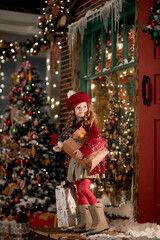 a little girl in warm clothes stands near the house with packages of New Year's gifts in her hands
