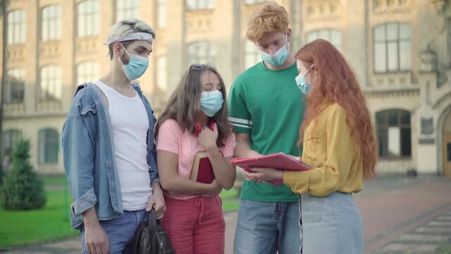 Four university students in face masks discussing homework project outdoors. Group of young Caucasian men and women standing on campus and talking during Covid-19 pandemic. Coronavirus education.
