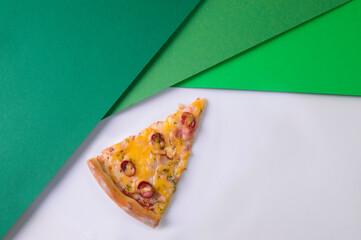 Close up of Cheesy slice of meat pizza with hunting sausages, cheese and basil pesto sauce on light background near colorful pieces of paper. Single piece of Italian pizza. Abstract green background
