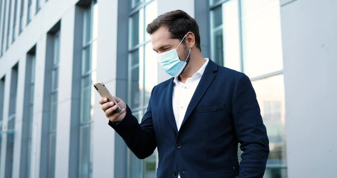 Caucasian young handsome man in medical mask tapping and scrolling on mobile phone outside at business center. Happy businessman in respiratory protection exting message on smartphone. Pandemic.