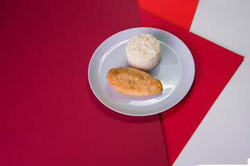 Side view of Tasty roasted chicken fillet on gray plate with boiled white rice served in culinary ring. Breakfast on colorful red background. Main and side dish in one plate.
