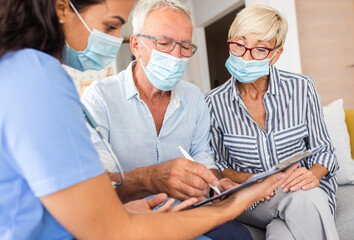 Female nurse talking to seniors patients with mask while being in a home visit, senior couple signs...