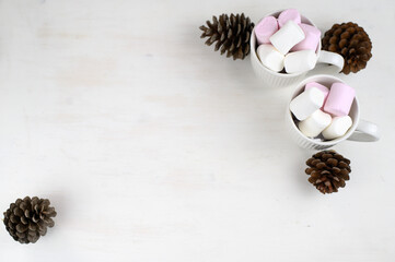 Hot winter drink with cocoa and marshmallows in mugs on wooden white background with Christmas garland and pine cones. Top view.