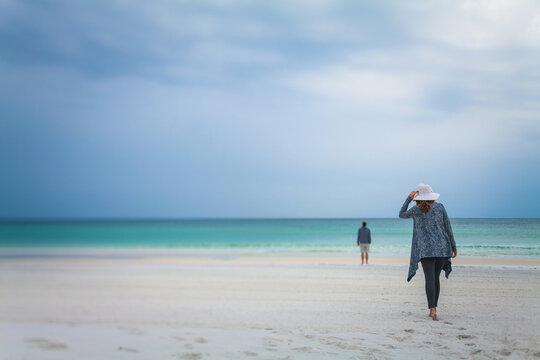 A woman wearing a hat on a white sandy beach walks towards a man standing at the edge of the sea. Ca