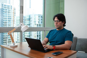 Young businessman with glasses dressed informal, working in his laptop in a wooden desk next to the window at home.