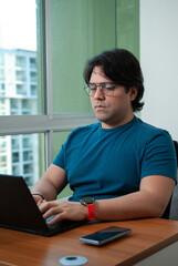 Vertical portrait of a young businessman with glasses dressed informal, working in his laptop next to the window in a wooden desk