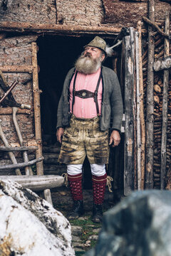 old bearded man in typical austrian outfit looking out of a wooden cabin