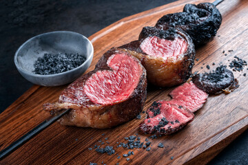 Modern style barbecue dry aged wagyu Brazilian picanha from the sirloin cap of rump beef sliced and offered as close-up on a skewer on a wooden design board on rustic black background