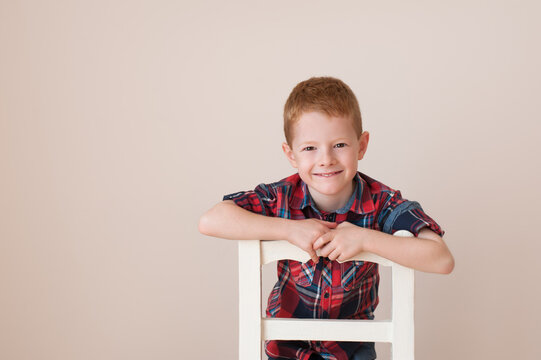 Portrait of an adorable redhead boy smiling, against a beige background kneeling on a white chair.