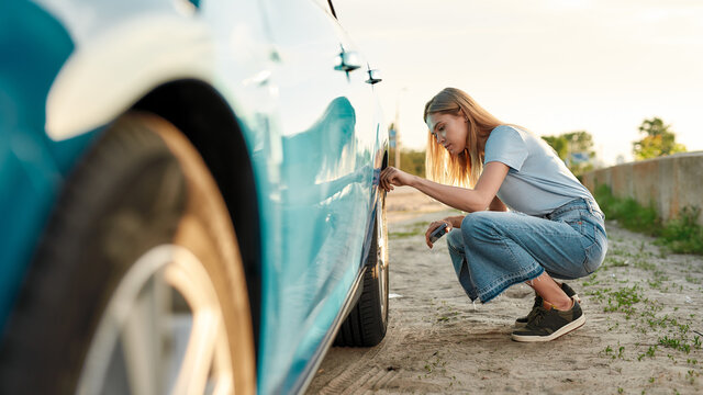 Attractive young woman checking air pressure of car tire on local road side while traveling