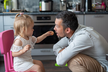 A cute little girl feeds cookies to her dad in the kitchen