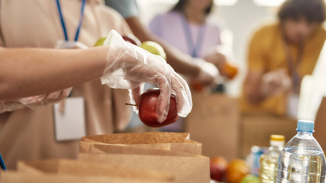 Close up of hand of volunteer in glove holding an apple while collecting, sorting food for needy people in paper bags, Team working together on donation project