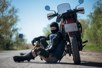 Tired biker is sitting on the road near his motorbike.
