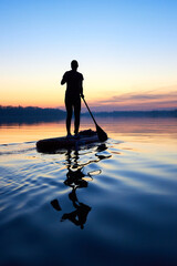 Silhouette of woman paddle on stand up paddle boarding (SUP) on quiet winter or autumn river at twilight. Calm river surface after sunset. Sports and meditation on the water