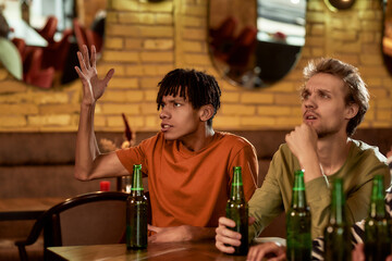 Young guy looking dissatisfied while watching sports match on TV, drinking beer and cheering for team in the bar. People, leisure and entertainment concept