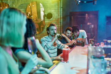 Three guys drinking beer, flirting, looking at women sitting at the bar counter. Friends spending time at night club, restaurant