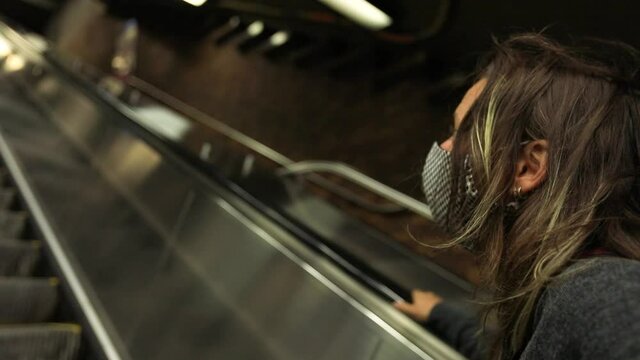 Portrait of young woman wearing coronavirus face protection mask standing with hand on rails going upstairs on escalator looking upwards