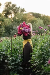 Deurstickers Very nice woman in a brown dress with a yellow apron, standing back to the camera at the dahlia farm, holding a bunch of freshly cut dahlia flowers in pink and purple color © anastasianess