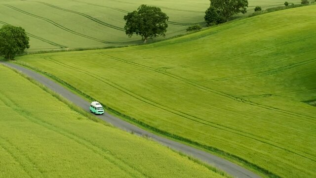 Mini bus driving on narrow country road, with green fields on both sides, drone stock footage 4
