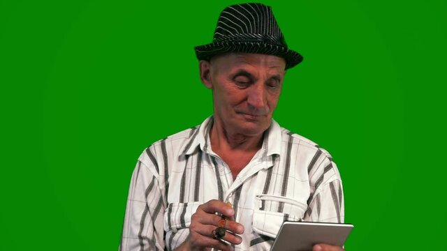 An elderly Caucasian man over 70 in an elegant hat with a smoking pipe in his lips has fun communicating on a digital gadget. Green screen