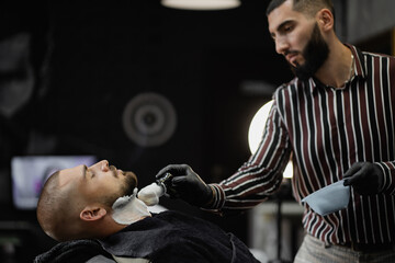 shaving foam is applied to the client's face, shaves the beard. beautiful hair, dark-haired young brutal man of European appearance is resting and waiting for the result.