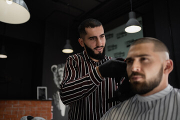 stylish men's haircut in barbershop short hair brunette Caucasian appearance. Beard and mustache on the face. Dark interior of the barbershop