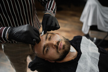 Fototapeta premium young man confident brutal Caucasian appearance, doing a haircut of his beard and hair on his head. dangerous razor blade shaving foam