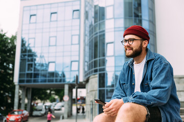 Cheerful bearded hipster male relaxed sitting in the city, holding smartphone, smiling and looking...