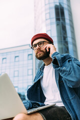 Serious stylish modern bearded entrepreneur wearing red hat and spectacles working on laptop and talking on phone