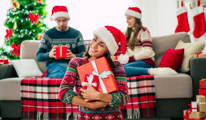 Merry Christmas and Happy New Year. Photo of a happy smiling beautiful woman in a Christmas Santa hat is showing a gift box in hands on group of friends and Christmas tree background.